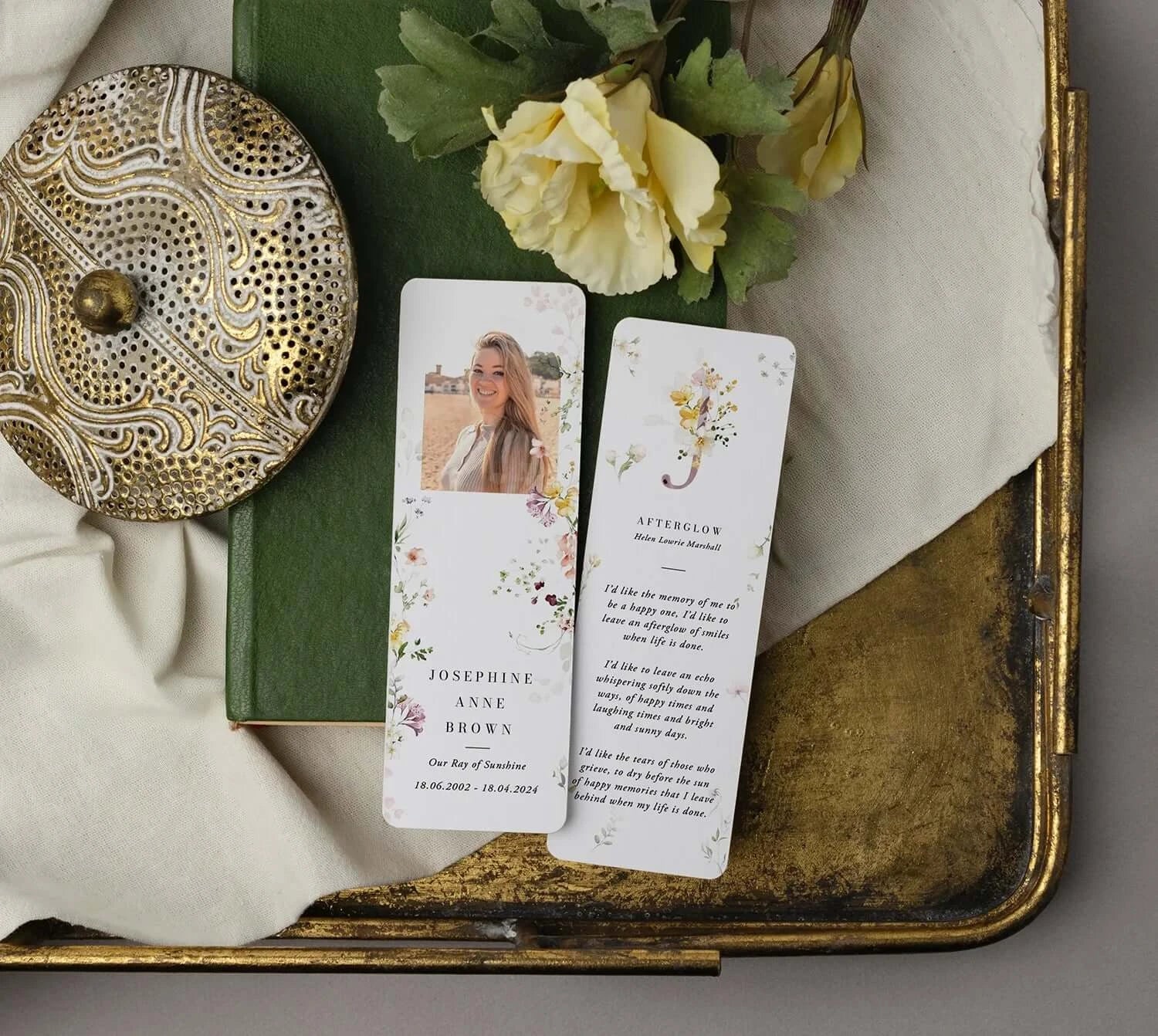 Memorial bookmark by Eternal Prints for Josephine Anne Brown, adorned with floral borders and her photo. Displayed with a vintage golden brooch and green book on cream fabric, symbolizing a cherished tribute